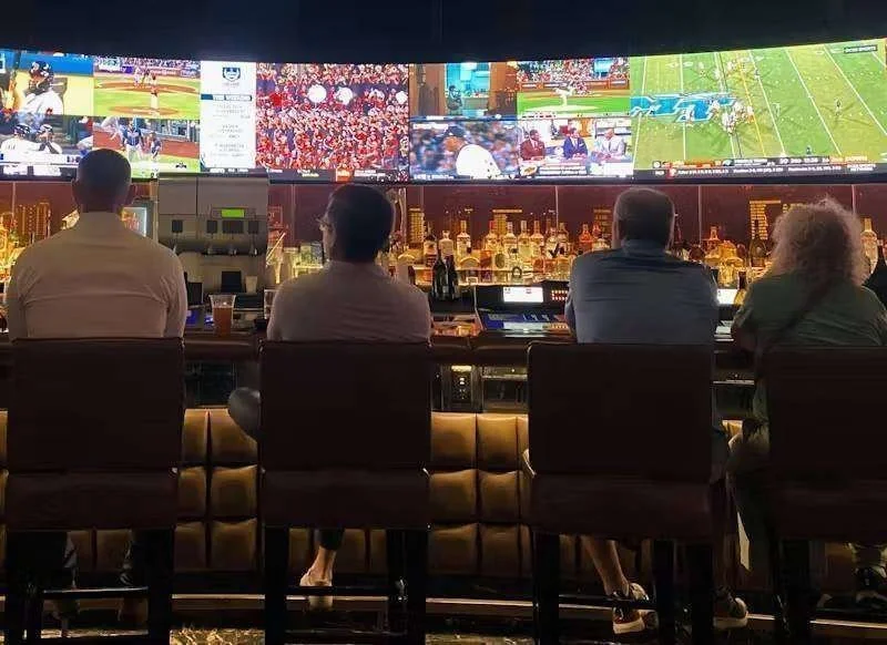 Quatro pessoas sentadas de costas em um bar, observando uma tela grande com diversas transmissões esportivas e garrafas de b…