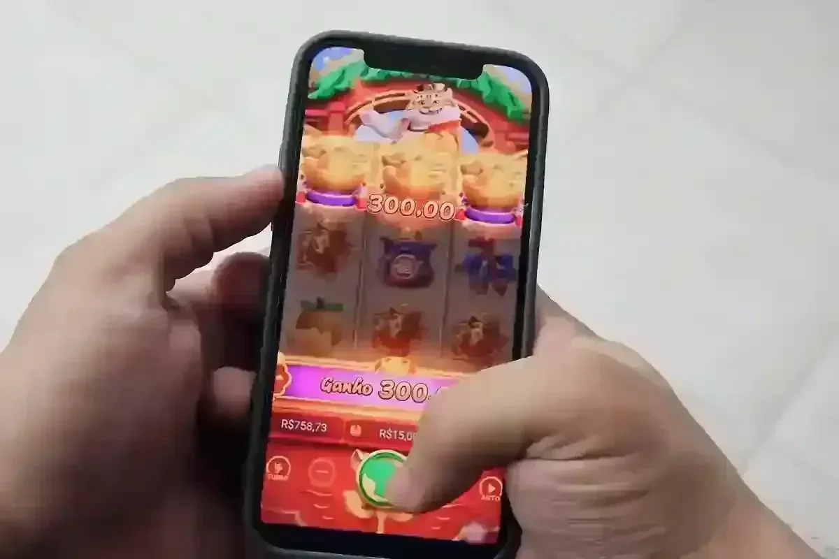 Mãos segurando um smartphone com jogo de caça-níqueis exibindo ganho de R$300,00 e saldo de R$758,73